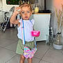toddler, child, indoor, floor_tiles, pigtails, white_top, green_shorts, black_shoes, pop_it_purse, butterfly_shaped, pink_container, curious_expression, living_room, tv_screen, high_chair, plant_decoration, glass_vase, mismatched_laces, socks, modern_interior