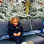 child, blue_eyes, pigtails, black_jacket, leggings, boots, bench, snow, pine_trees, frost, deer_statue, antlers, winter, outdoor, smiling, curly_hair, holiday_decor, playful, park, daytime