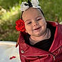 baby, child, face, smile, red_jacket, flower, headband, outdoor, greenery, portrait, cute, happy, infant, fashion, closeup, baby_clothing, head_accessory, nature, soft_light, sitting