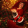 woman, cat, holiday, christmas, festive_clothing, plaid_blanket, red_chair, glasses, smile, indoor, decorations, lights, greenery, cozy, pet, santa_outfit, relaxing, warm_lighting, celebration, portrait