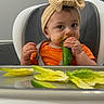 baby, child, high_chair, headband, cucumber, lettuce, food, messy_face, orange_shirt, indoor, feeding, person, cute, eating, toddler, kitchen, healthy_food, infant, plate, seat