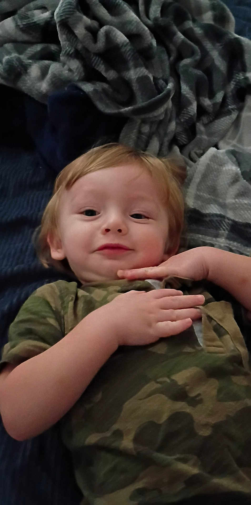 Jace is registered to the contest to win money with this photo: toddler, child, smiling, face, hands, camouflage_clothing, blanket, fabric, soft_texture, lying_down, indoor, portrait, cute, young_child, casual_clothing, relaxed, resting, head, person, skin