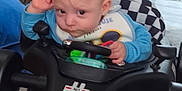 Martin is registered to the contest to win money with this photo: baby, child, indoor, toy_car, steering_wheel, blue_clothing, bib, car_seat, car_toy, floor, carpet, face, hand, footwear, sock, hair, expression, furniture, window, light