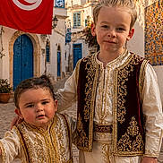 Nëlya Saidi a rejoint le concours — aidez-le/la à gagner de superbes lots ! child, children, traditional_clothing, tunisian_flag, street, cobblestone, architecture, building, door, window, flower, foliage, sky, outdoor, portrait, smiling, people, culture, costume, town