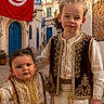 child, children, traditional_clothing, tunisian_flag, street, cobblestone, architecture, building, door, window, flower, foliage, sky, outdoor, portrait, smiling, people, culture, costume, town
