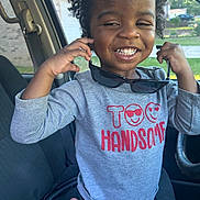 Kainen is registered to the contest to win money with this photo: car_interior, casual_clothing, child, curly_hair, cute, daylight, grey_shirt, hand_raised, happy, indoors, joyful, person, portrait, red_text, seat, seatbelt, smiling, sunglasses, window, young_child