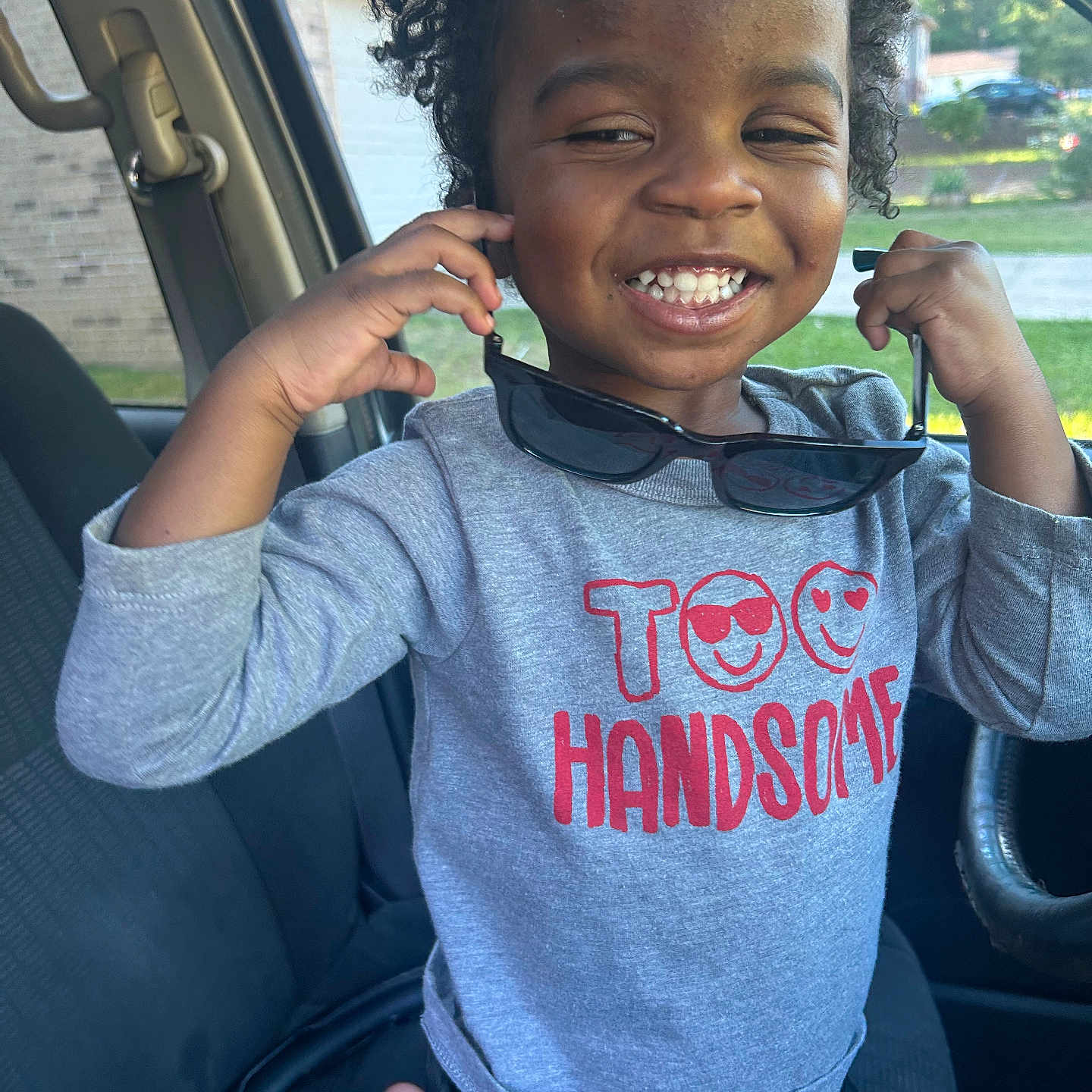 Kainen is registered to the contest to win money with this photo: car_interior, casual_clothing, child, curly_hair, cute, daylight, grey_shirt, hand_raised, happy, indoors, joyful, person, portrait, red_text, seat, seatbelt, smiling, sunglasses, window, young_child