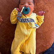 Aziel joined the competition — help win amazing prizes! baby, pacifier, yellow_onesie, infant, lying_down, hand, face, soft_surface, brown_background, clip, cozy, newborn, cute, small, peaceful, child, indoors, sleepwear, person, portrait