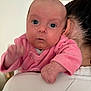 baby, infant, pink_clothing, person, holding, shoulder, face, hand, blue_eyes, indoor, blurred_hand, closeup, expression, head, hair, adult, skin, portrait, young, candid