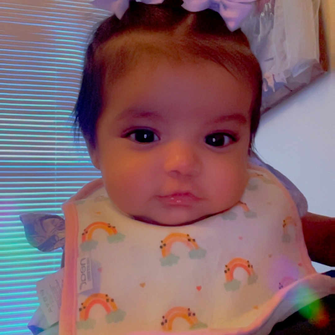 Feliciana is registered to the contest to win money with this photo: baby, child, bib, bows, hair, indoor, person, portrait, cute, face, hand, clothing, decor, window_blinds, red_hat, rainbow_pattern, infant, skin, smile, holding