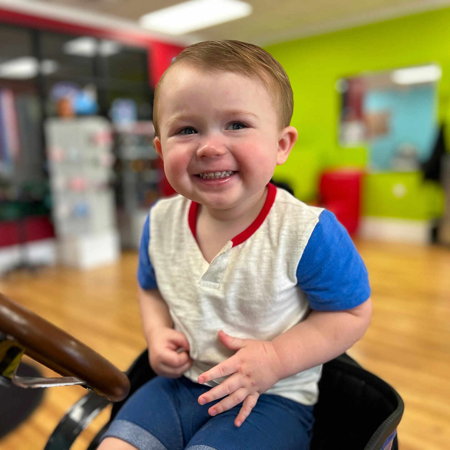 Bryson is registered to the contest to win money with this photo: toddler, child, smiling, portrait, stroller, indoor, colorful, playroom, face, person, blue_shirt, white_shirt, short_hair, happy, sitting, wooden_floor, background, baby, teeth, candid
