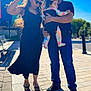 family, baby, man, woman, outdoor, daylight, sunglasses, dress, jeans, pavement, bench, street, holding, child, sunlight, shadow, urban, happy, people, colorful_clothing