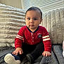 baby, child, red_sweater, black_pants, soft_toys, cushions, window_blinds, gray_quilt, indoor, seated, curious_expression, natural_light, hand, footwear, plush, portrait, cozy, home, cute, young_child