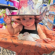 Lillyth is registered to the contest to win money with this photo: aisle, baby, black_tank_top, cart, child, colorful, cowboy_hat, elephant_toy, floral_pattern, hat, indoor, infant, playful, plush_toy, portrait, retail_store, shopping, shopping_cart, smiling, supermarket