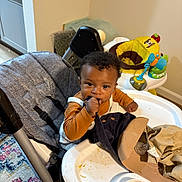 Isaiah joined the competition — help win amazing prizes! baby, child, curly_hair, expression, fabric, face, feeding, floor_rug, high_chair, home, indoor, infant, messy, orange_shirt, person, plastic_bowl, play_area, seatbelt, table, toy