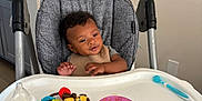 Isaiah is registered to the contest to win money with this photo: baby, bib, bottle, child, curly_hair, feeding, happy, high_chair, indoor, infant, kitchen, messy, person, plastic_bowl, seat, smiling, spoon, table, toy, young_child