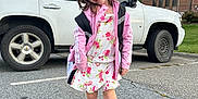 Sophia is registered to the contest to win money with this photo: child, girl, smiling, posing, parking_lot, white_suv, backpack, pink_jacket, floral_dress, sneakers, outdoor, asphalt, building, cloudy_sky, trees, casual_clothing, happy, playful, daylight, sidewalk