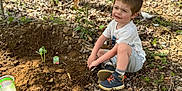 Killian joined the competition — help win amazing prizes! child, boy, outdoor, nature, planting, soil, seedling, watermelon, garden, dirt, boots, white_tshirt, shorts, sunlight, leaves, woods, young_child, playing, ground, happy