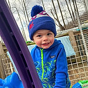 Killian is registered to the contest to win money with this photo: toddler, child, blue_jacket, hat, pom_pom, smile, outdoor, playground, toy_truck, fence, trees, cloudy_sky, wooden_deck, play, cold_weather, happy, person, clothing, hand, face