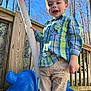 child, boy, smiling, plaid_shirt, blue_shirt, green_shirt, tan_pants, outdoor, playground_toy, blue_toy, wooden_deck, pole, trees, sunny, daylight, happy, person, play, nature, casual_clothing