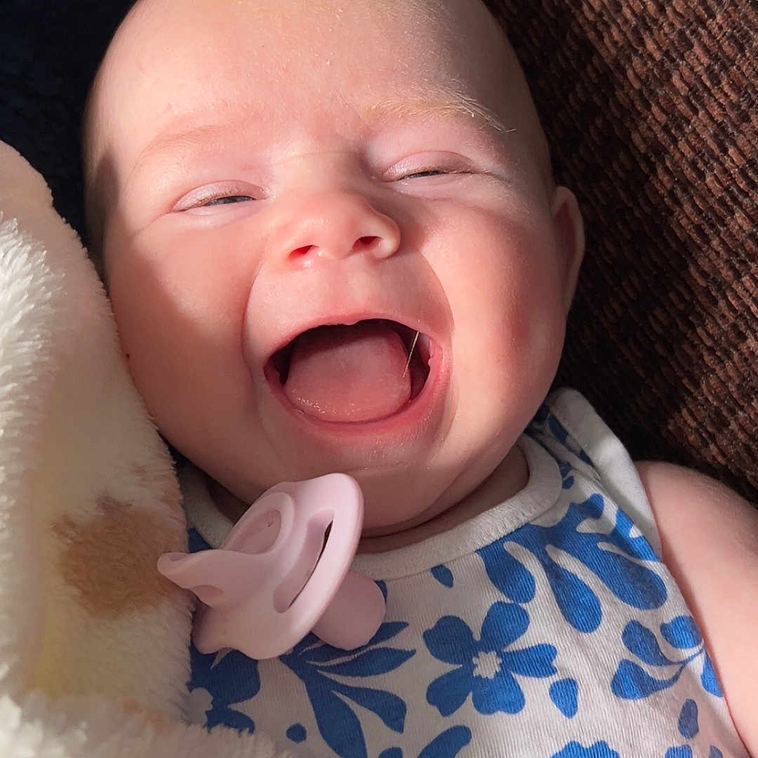 Amelia is registered to the contest to win money with this photo: baby, blanket, blue_flower_pattern, child, closeup, comfort, cozy, cute, expression, face, happy, indoor, infant, light, pacifier, portrait, skin, sleepwear, smiling, soft_texture