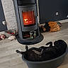 dog, dogs, pet, black_dog, sleeping, lying_down, upside_down, relaxed, pet_bed, indoor, living_room, wood_stove, fire, warmth, cozy, wood_floor, home, furniture, animal, resting
