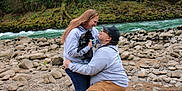 Koda is registered to the contest to win money with this photo: river, rocks, outdoor, nature, greenery, trees, person, dog, black_dog, kneeling, hugging, casual_clothing, sneakers, happy, affection, companionship, smiling, shorts, hoodie, cap