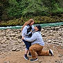 river, rocks, outdoor, nature, greenery, trees, person, dog, black_dog, kneeling, hugging, casual_clothing, sneakers, happy, affection, companionship, smiling, shorts, hoodie, cap