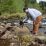 Koda joined the competition — help win amazing prizes! person, dog, rock, stream, water, forest, nature, outdoor, greenery, cloudy_sky, casual_clothing, hat, glasses, petting, small_dog, bending, rocks, moss, gravel, trees