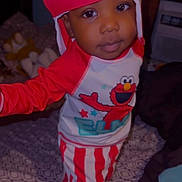 Amias is registered to the contest to win money with this photo: baby, child, infant, portrait, standing, costume, red_hat, striped_pants, cartoon, character, elmo, smiling, indoor, bedroom, bed, patterned_blanket, fashion, cute, face, photography