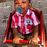 Amias is registered to the contest to win money with this photo: child, toddler, baby, portrait, sitting, high_chair, smiling, pacifier, red_shirt, plaid, white_shoes, toy, earring, outdoor, sunlight, brick_wall, stone_pavement, furniture, happy, front_view
