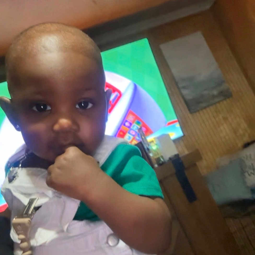 Amias joined the competition — help win amazing prizes! child, toddler, baby, portrait, sitting, indoors, face, eyes, hand, smiling, curious, clothing, overalls, green_shirt, television, living_room, furniture, wood_paneling, photograph, home