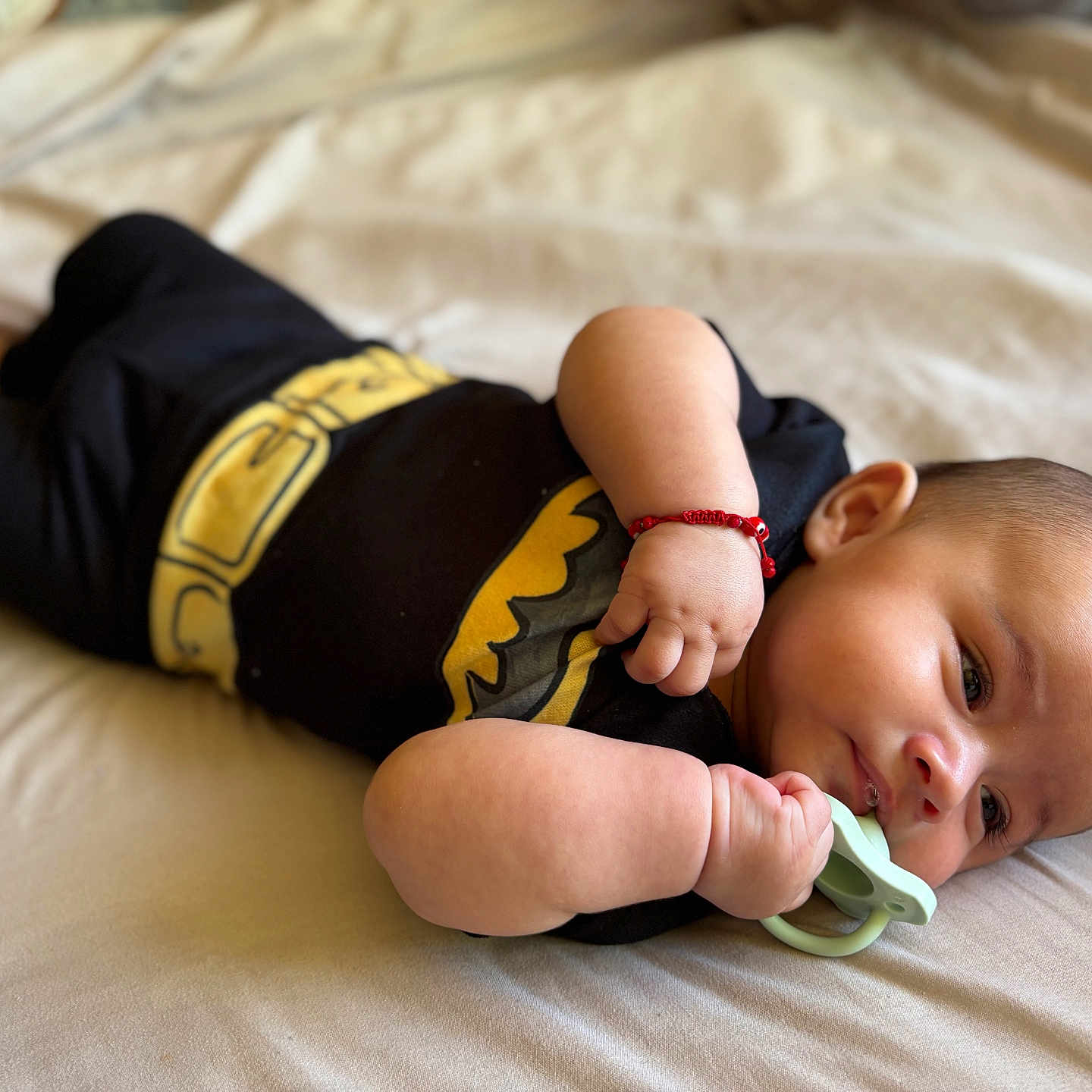 Agustin is registered to the contest to win money with this photo: baby, infant, pacifier, batman_outfit, lying_down, bed, bracelet, cute, child, skin, hand, face, head, clothing, young, resting, indoors, sleepwear, adorable, portrait