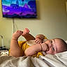 baby, infant, bed, blanket, yellow_clothing, hand, foot, face, lying_down, indoors, television, blurred_background, cute, chubby, skin, person, resting, sleepy, red_bracelet, cozy