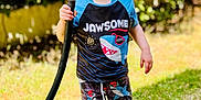 Jameson is registered to the contest to win money with this photo: child, smiling, happy, grass, outdoor, barefoot, hose, water, wet_hair, shirt, shorts, summer, sunny, nature, greenery, playful, cute, person, toddler, daylight
