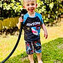 child, smiling, happy, grass, outdoor, barefoot, hose, water, wet_hair, shirt, shorts, summer, sunny, nature, greenery, playful, cute, person, toddler, daylight