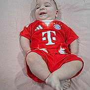Asher joined the competition — help win amazing prizes! baby, child, infant, lying_down, red_clothing, sports_jersey, football_kit, smiling, happy, cute, indoor, soft_surface, short_hair, person, young_child, casual, relaxed, comfortable, portrait, single_person