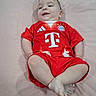 baby, child, infant, lying_down, red_clothing, sports_jersey, football_kit, smiling, happy, cute, indoor, soft_surface, short_hair, person, young_child, casual, relaxed, comfortable, portrait, single_person