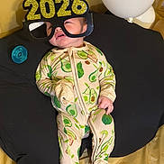 Alexander is registered to the contest to win money with this photo: baby, infant, child, pajamas, avocado_pattern, glasses, novelty_glasses, 2026, balloon, pacifier, cushion, black_cushion, yellow_background, footed_pajamas, sleepwear, indoors, cute, expression, smiling, comfort