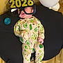 baby, infant, child, pajamas, avocado_pattern, glasses, novelty_glasses, 2026, balloon, pacifier, cushion, black_cushion, yellow_background, footed_pajamas, sleepwear, indoors, cute, expression, smiling, comfort