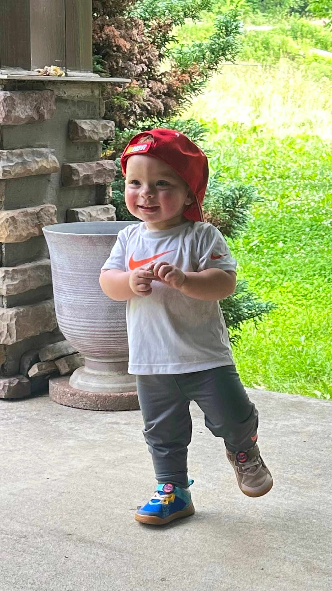 Chad is registered to the contest to win money with this photo: toddler, child, red_cap, white_tshirt, gray_pants, sneakers, outdoor, porch, stone_wall, greenery, plant_pot, smiling, walking, concrete_floor, daylight, casual_clothing, happy, cute, nature, playful
