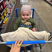 Liam is registered to the contest to win money with this photo: toddler, shopping_cart, grocery_store, child, blanket, hat, smile, hand, ring, aisle, shelves, products, floor, clothing, jeans, socks, metal, wire_cart, person, indoor