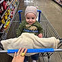 toddler, shopping_cart, grocery_store, child, blanket, hat, smile, hand, ring, aisle, shelves, products, floor, clothing, jeans, socks, metal, wire_cart, person, indoor