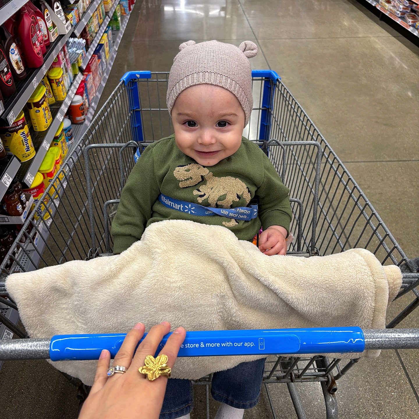 Liam is registered to the contest to win money with this photo: aisle, blanket, child, clothing, floor, grocery_store, hand, hat, indoor, jeans, metal, person, products, ring, shelves, shopping_cart, smile, socks, toddler, wire_cart