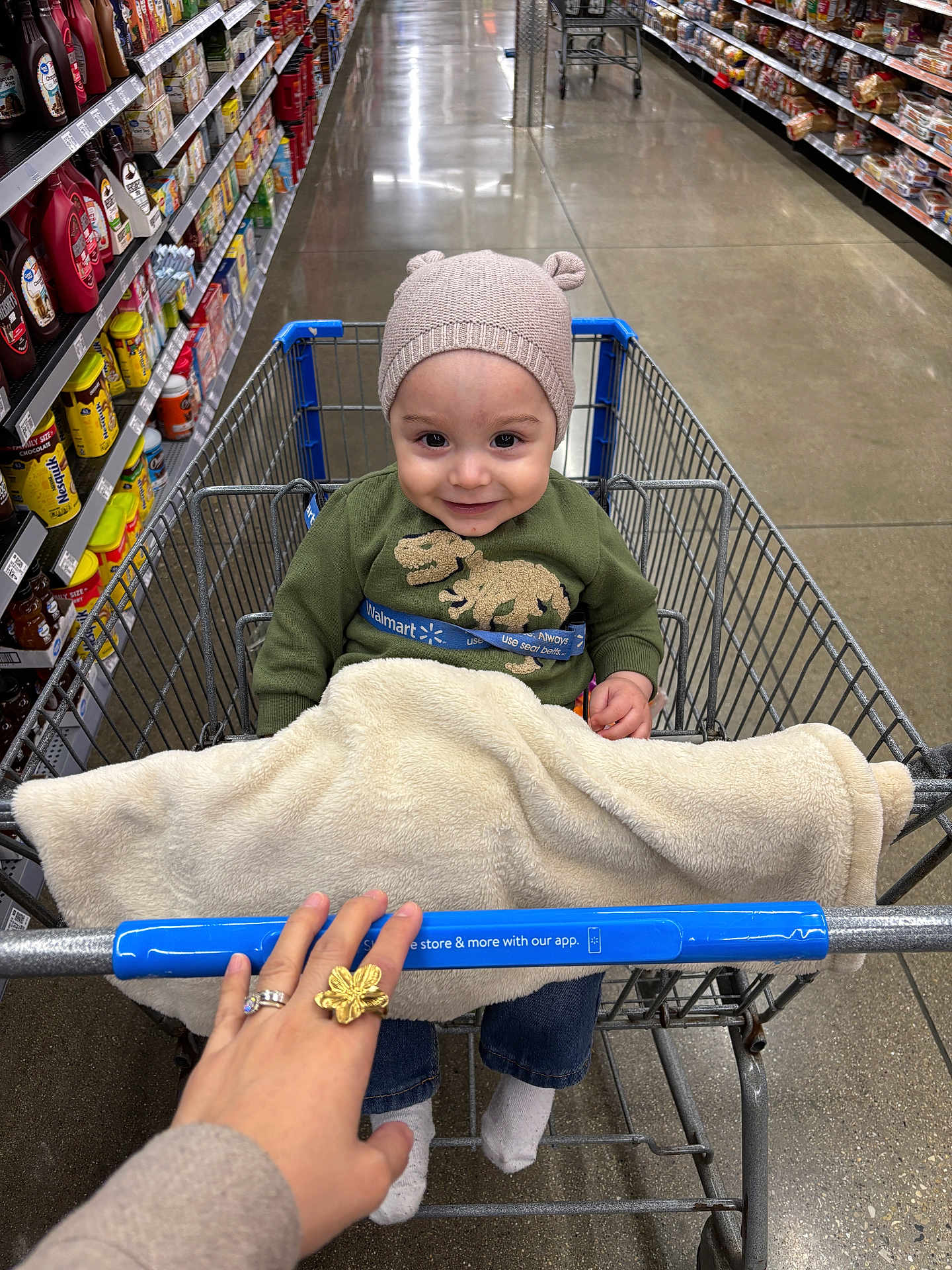 Liam is registered to the contest to win money with this photo: toddler, shopping_cart, grocery_store, child, blanket, hat, smile, hand, ring, aisle, shelves, products, floor, clothing, jeans, socks, metal, wire_cart, person, indoor
