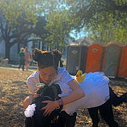 Archie is registered to the contest to win money with this photo: woman, dog, tutu, rubber_duck, hugging, outdoor, sunlight, trees, grass, people, portable_toilets, park, costume, black_dog, candid, affection, casual, daytime, smiling, fun