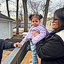 Amalia is registered to the contest to win money with this photo: child, woman, person, outdoor, jacket, glasses, hand, wooden_bench, railing, trees, house, winter, cloudy_sky, footwear, jeans, playful, family, support, smile, pigtails