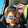 child, car_seat, sunglasses, curly_hair, sunlight, car_interior, seatbelt, window, reflection, black_clothing, road_trip, portrait, young_child, casual, travel, seat, safety_harness, daylight, cute, relaxed