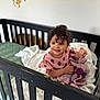 toddler, crib, blanket, curly_hair, smiling, child, indoor, bed, bedding, knitted_blanket, pink_clothing, wall_decor, lion_decal, monkey_decal, happy, person, baby, cute, nursery, furniture