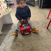 Azariah is registered to the contest to win money with this photo: toddler, child, toy_car, indoor, carpet, curly_hair, hoodie, pants, boots, crib, play_toy, table, furniture, couch, blanket, carpeted_floor, room, person, curious_expression, steering_wheel